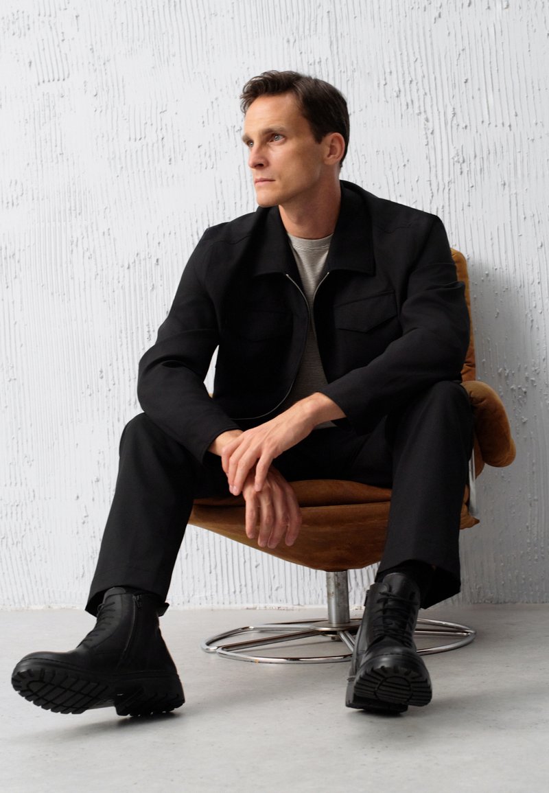 Czarna kurtka z dwoma przednimi kieszeniami, paskowana koszula pod spodem, czarne spodnie i masywne czarne buty. Siedzi na okrągłym brązowym krześle.