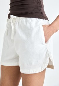 Shorts en coton blancs avec un cordon de serrage à la taille, des poches latérales et un ourlet légèrement arrondi. Tissu léger avec une texture lisse.