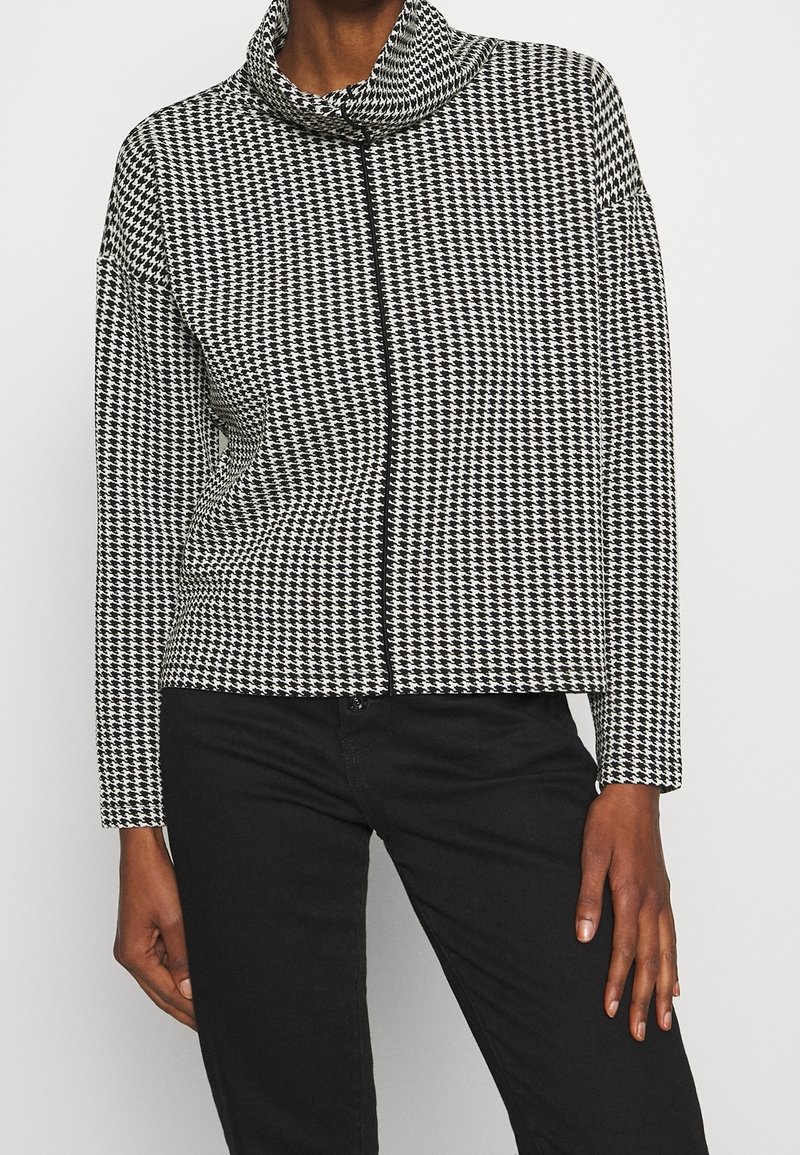 Femme portant un haut à manches longues à motif pied-de-poule noir et blanc avec un col montant et un pantalon noir, debout devant un fond uni.