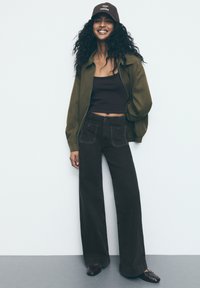 Dunkelblaue weite Jeanshose mit übergroßen Taschen, kombiniert mit einem enganliegenden schwarzen Crop-Top und einer olivgrünen Bomberjacke. Braune Kappe mit Text.