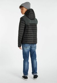 Giubbotto imbottito nero con pannello in fleece verde sulla spalla, dotato di cappuccio e texture a coste. Indossato con jeans blu e sneaker nere.