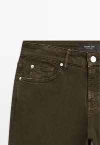 Jean en denim vert foncé avec une texture légère, dotés d'un design à cinq poches, de finitions en métal argenté et d'une étiquette sur la taille.