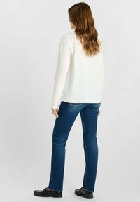 Witte blouse met lange mouwen, gecombineerd met donkerblauwe rechte jeans; het model draagt zwarte instappers en heeft golvend haar, gezien vanuit de achterkant.