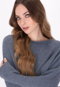 Pull en tricot gris avec un motif texturé, coupe ample et encolure large. Cheveux longs et ondulés encadrant le visage, éclairage doux.