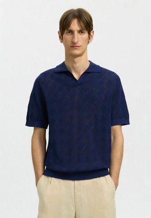 Homme aux cheveux bruns courts portant un polo bleu marine texturé à manches courtes et un pantalon beige, debout les mains dans les poches.