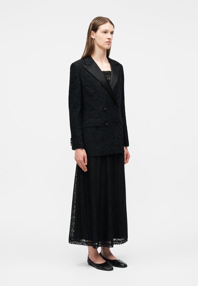 Γυναίκα που φοράει μακρύ μαύρο δαντελένιο φόρεμα με υφασμένο μαύρο διπλό σακάκι και μαύρες μπαλαρίνες, στέκεται μπροστά σε ένα απλό λευκό φόντο.