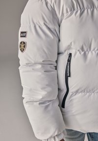 Giacca a piumino bianca con design trapuntato, con zip nera e toppe con logo. La giacca ha polsini elastici e una texture liscia e lucida.