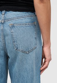 Δεξί χέρι ακουμπισμένο δίπλα σε ανοιχτό μπλε τζιν με ορατή πίσω τσέπη και μαύρο υφασμάτινο επάνω άκρο.