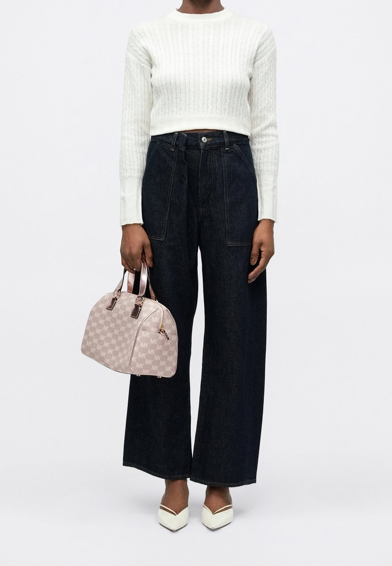 Femme portant un pull court en maille torsadée blanche, un jean large foncé, des chaussures blanches pointues, tenant un sac à main beige à motifs.