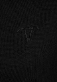 Schwarzer Strickstoff mit einem geprägten Stierkopf-Logo in der Mitte, das einen subtilen Textur-Kontrast zum glatten Hintergrund schafft.