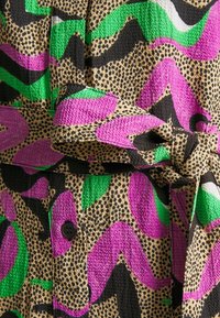 Tissu texturé avec un motif de points noirs sur beige, surmonté de formes abstraites vert vif, rose et noir, et un détail de ceinture nouée.