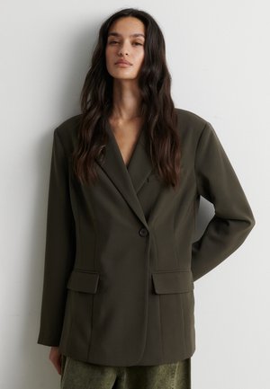 Donna con i capelli lunghi e scuri che indossa un blazer verde scuro oversize e pantaloni verdi, in piedi contro una parete chiara e liscia.