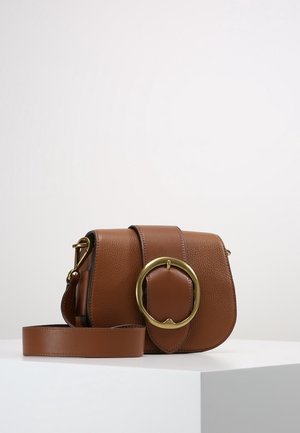 Bruine leren crossbodytas met grote ronde gouden gesp en verstelbare riem, geplaatst op een witte ondergrond tegen een effen achtergrond.