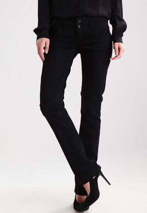 Kvinde iført sort slim-fit jeans og sorte højhælede sko, stående med krydsede ben mod en ensfarvet baggrund.