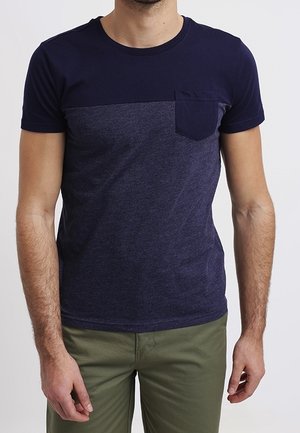 Homme portant un t-shirt à manches courtes bicolore bleu marine et bleu foncé avec une poche poitrine et un pantalon vert olive.