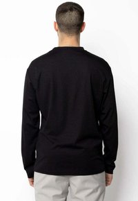 Homme à cheveux courts portant une chemise à manches longues noire et un pantalon gris clair, se tenant face à un mur blanc avec les mains détendues sur les côtés.