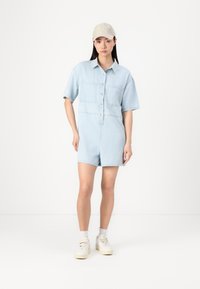 Svijetloplav denim kombinezon kratkih rukava s kopčanjem sprijeda, dva džepa na prsima i opuštenim krojem, u paru s bež kapom i bijelim tenisicama.