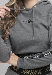 Grijze hoodie met trekkoord en gouden beslag, voorzien van een zwart logo. Band eronder met gouden accenten en merknaam in vetgedrukte letters.
