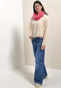 Giubbotto beige corto, maglione leggero, jeans a gamba larga blu, sciarpa rosa a motivo. Outfit casual con texture morbide e colori a contrasto.