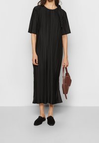 Schwarzes Faltenkleid mit kurzen Ärmeln, rundem Ausschnitt und einer Schleifendetail an der Seite. Kombiniert mit schwarzen Slip-On-Schuhen und einer braunen Handtasche.