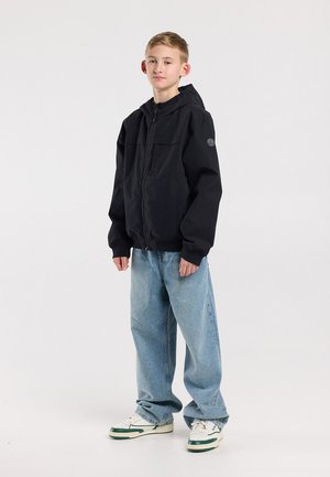 Tienerjongen staand, gekleed in een zwarte capuchonjas, wijde lichtblauwe spijkerbroek en witte sneakers met groene accenten, tegen een effen lichte achtergrond.