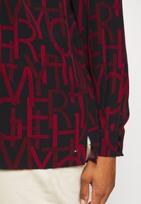 Zwarte shirt met een gedurfde, rode grafische patroon van letters. Inclusief knoopsluitingen bij de mouwen en zijsplitten voor extra stijl. Zachte stofstructuur.