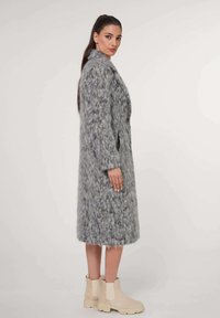 Langer grauer Texturobermantel aus einem weichen, flauschigen Material, mit einem Knopfverschluss und seitlichen Taschen, kombiniert mit beigen Ankle Boots.