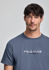 Grå t-shirt med kort ärm och rund halsringning, med vit text "POLO CLUB" och "ETABLISHED 2012" i mitten. Slät textur.