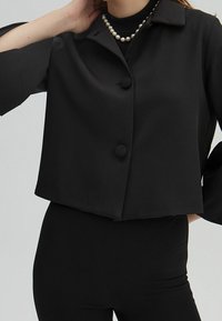 Svart kort jacka gjord av slät tyg, med tre knappar, spetsig krage och vida ärmar. Bärs över en tajt svart topp.