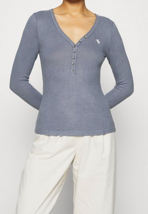 Person wearing a fitted grey long-sleeve henley shirt with white logo on chest, paired with loose white pants against a plain background.