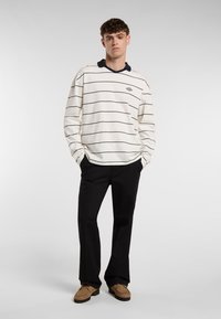 Langærmet, creme-stribet polo med navy krave, parret med sorte bukser og brune snørebåndssko, stående mod en neutral baggrund.