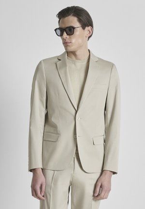 Mann trägt beigen Anzug und passenden Pullover mit schwarzer Sonnenbrille, steht vor einfachem grauem Hintergrund.