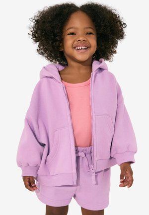 Smilende ung pige med krøllet hår iført en lyserød ribbet top under en lys lilla hættetrøje med lynlås og matchende shorts.