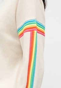 Πλεκτό πουλόβερ με μπεζ ύφασμα. Διαθέτει πολύχρωμη ρίγα ουράνιου τόξου κατά μήκος του μανικιού σε αποχρώσεις ροζ, πορτοκαλί, πράσινο και μπλε.