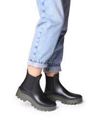 Botas de tobillo de goma negra con paneles laterales elásticos, suelas gruesas verdes y detalles texturizados. Combinadas con jeans claros de corte acampanado.