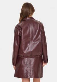 Donna con lunghi capelli castani che indossa una giacca in pelle sintetica di un bordeaux scuro e una gonna abbinata, vista di spalle contro uno sfondo bianco.