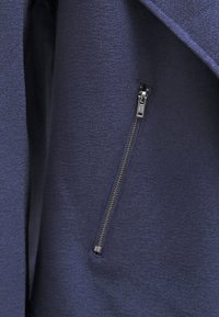 Tissu bleu marine avec une finition texturée, comportant une fermeture éclair argentée sur le côté gauche, intégrée dans un design de veste.