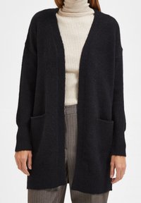 Mörk svart cardigan i mjukt, texturerat material, med avslappnad passform, öppen front och två framfickor.