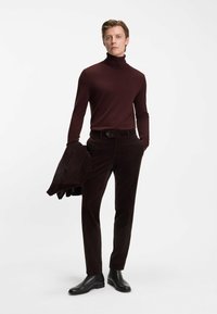 Bordeaux turtleneck, donkerbruine corduroy broek en zwarte leren enkellaarsjes. Model houdt een bijpassende jas over de linkerarm.