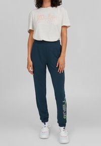 Svetle šedé tričko s multibarevným nápisem 'O'Neill California', spárované s tmavě modrými tepláky s potiskem zeleného a bílého loga. Bílé tenisky.