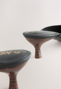 Schwarze Lederschuhe mit Absatz und geschwungenen Holzabsätzen. Die Innensohlen zeigen dezente Nähte und ein goldenes Logo und präsentieren ein elegantes, modernes Design.