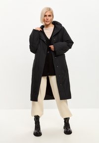 Sort hættepuffercoat med store knapper, quiltet tekstur og sidelommer, båret over en mørk cardigan og cremefarvede bukser.
