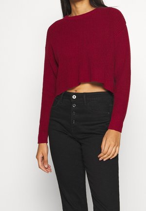 Pull court en maille côtelée rouge avec manches longues, associé à un jean noir taille haute avec un devant à boutons.