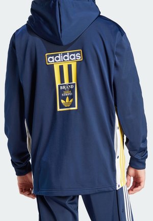 Personne portant une veste Adidas bleu marine avec des bandes jaunes et blanches et un patch logo Adidas dans le dos, debout dos tourné vers l'avant.