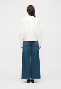 Λευκό φούτερ με στρογγυλή λαιμόκοψη και χαλαρή ραφή στους ώμους, συνδυασμένο με φαρδιά μπλε τζιν παντελόνια. Το τζιν διαθέτει πίσω τσέπες και άκοπο τελείωμα.