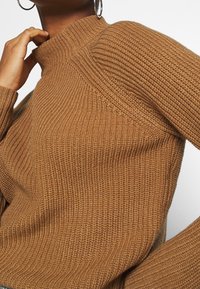 Personne portant un pull à col roulé marron côtelé, touchant son cou, avec une grande boucle d'oreille argentée visible et visage partiellement cadré.