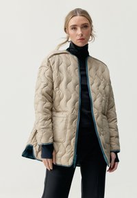 Giacca beige trapuntata con chiusura a zip, con accenti turchesi e vestibilità rilassata. Include tasche laterali e un colletto alto per maggiore calore.