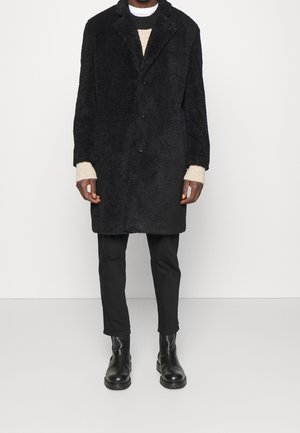 Mężczyzna w czarnym teksturowanym płaszczu do kolan, beżowym swetrze, czarnych spodniach i czarnych skórzanych butach, stojący na jednolitym tle.