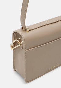 Sac à main rectangulaire beige avec des ferrures dorées et une poignée supérieure, arborant le logo Steve Madden embossé dans le coin inférieur avant.