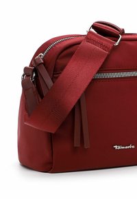 Red fabric crossbody bag with silver hardware, adjustable strap, zip closure, and side tassels. Logo is embossed on the front.